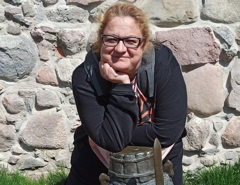 Kobieta w wieku średnim, jasne lekko kręcone włosy zebrane w kucyk, ubrana na czarno. Za nią kamienna ściana. Kobieta opiera się o głowę posągu krasnala. Krasnal na głowie ma hełm z dwoma rogami, a w ręku uniesiony miecz.