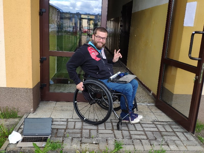 Mężczyzna w średnim wieku na wózku Prawa dłoń umieszczona na ciągu, lewa nieznacznie uniesiona w geście zwycięstwa. Za mężczyzną otwarte widoczne szklane drzwi w brązowej oprawie. Za nimi korytarz od podłogi do połowy pomalowany na żółto, reszta ścian biała. Na chodniku po prawej stoi otwarty laptop.