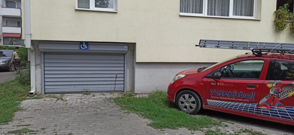 Widok na żółta ścianę bloku, widoczne okna na parterze budynku. Po lewej stronie budynku widoczna srebrna , podnoszona do góry brama wjazdowa do garażu. Na górnej framudze bramy widoczna kwadratowa tabliczka- na niebieskim tle biały symbol osoby na wózku. Brama jest bardzo niska. Dla porównania po prawej stronie kadru czerwony samochód osobowy z drabiną na dachu - samochód jest wyższy niż wjazd do garażu. Samochód ma kolor czerwony a na nim niebieską czcionką umieszczony jest napis TeleKabel i umieszczona grafika ludzika trzymającego wielka klawiaturę pod pachą.