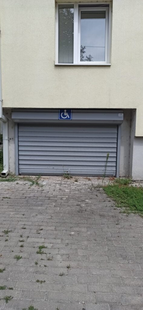 Widok na żółta ścianę bloku, widoczne okna na parterze budynku. Po lewej stronie budynku widoczna srebrna , podnoszona do góry brama wjazdowa do garażu. Na górnej framudze bramy widoczna kwadratowa tabliczka- na niebieskim tle biały symbol osoby na wózku. Brama jest bardzo niska.