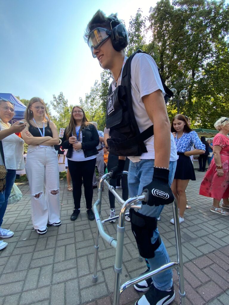 Słoneczny dzień, niebieskie niebo, deptak w parku. Na deptaku stoją w rozsypce ludzie ubrani w letnie ubrania. Na pierwszym planie młody mężczyzna ubrany w symulator starości przemieszcza się przy wsparciu balkonika. Nastrój zdjęcia pogodny.