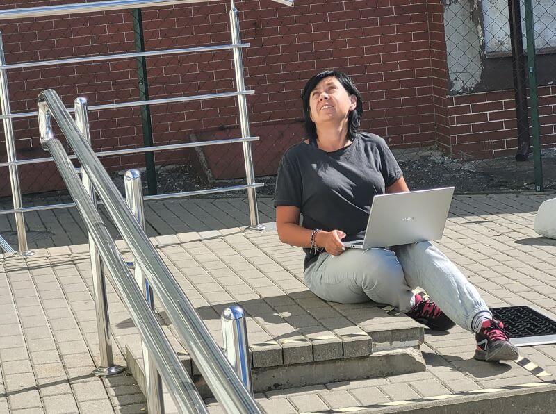 Widok na schody zewnętrzne i kawałek pochylni. Na górnym podeście schodów siedzi brunetka w wieku średnim. Ubrana jest w jasnoniebieskie spodnie i ciemnoszarą bluzkę, na nogach buty sportowe czarne z różowymi wstawkami. Kobieta odrzuca głowę do tyłu – sprawdza czy nad niewidocznym na zdjęciu drzwiami wejściowymi jest daszek. Na kolanach otwarty laptop.