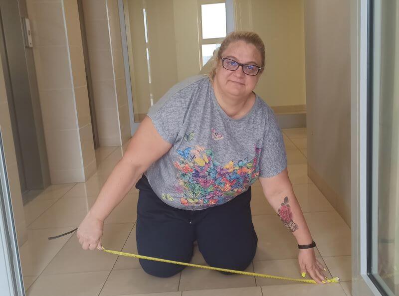 Jasne wnętrze. Kobieta w wieku średnim, jasne lekko kręcone włosy zebrane w kucyk, ubrana w szarą bluzkę z motywem odlatujących kolorowych motyli i ciemne spodnie. Kobieta klęczy i dokonuje pomiaru szerokości drzwi. W dłoniach trzyma rozciągniętą miarkę. Na lewej ręce kobiety widoczny duży tatuaż – wzór kwiatowy.