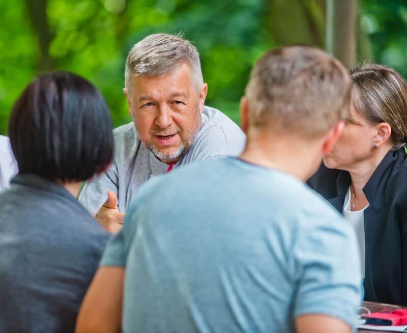Trzy osoby siedzą przy niewidocznym na zdjęciu stole – dwie tyłem do osoby robiącej zdjęcie i jedna na przeciw nich patrząca w skupieniu na jedną z tych osób. Na zdjęciu rozmazane tyły głów osób siedzących tyłem do osoby fotografującej. Między głowami widoczna twarz mężczyzny w wieku średnim uchwycona w czasie jego wypowiedzi. Mężczyzna ma jasne włosy, a na twarzy siwy zarost.