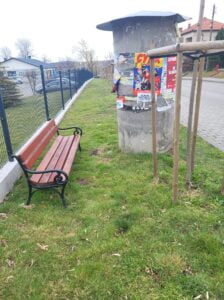 Trawnik towarzyszący chodnikowi. Na trawniku bokiem do kadru stoi drewniana ławka z podłokietnikami i oparciem. Ławka ustawiona jest w głębi trawnika tuż przy płocie otaczającym jakąś posesje. Na wprost siedziska ławki betonowy, okrągły słup ogłoszeniowy i młode drzewko otoczone 3 palami. Odległość od ławki do opisanych elementów niewielka co utrudnia dotarcie do siedziska.