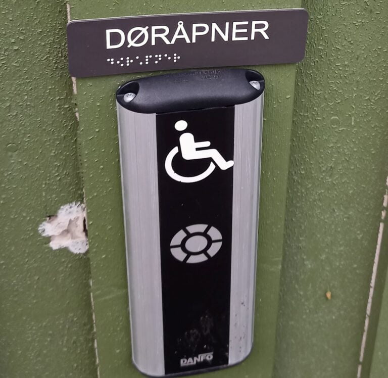 Na zielonej ścianie czarno-szary element – przycisk otwierania drzwi lub przywoływania pomocy. Nad nim napis Dorapner (napis po norwesku) oraz napis w alfabecie Braille’a. Napis wykonany dużą, białą czcionką znajduje się na ciemny tle, również litery a alfabecie Braille’a są jasne i umieszczone na tej samej ciemnej tabliczce. Sam przycisk ma postać prostopadłościanu a jasny długich krawędziach. Na jego czarnej środkowej części znajdują się jasne symbol osoby poruszającej się na wózku i koło symbolizujące przycisk. Symbol osoby na wózku jest podświetlony.
