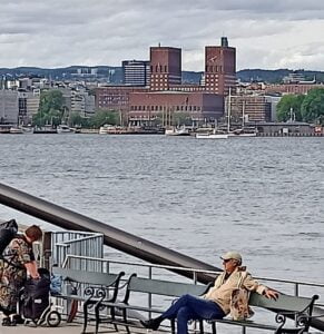 Widok na Oslo od strony fiordu. W tle ratusz miejski. Po stronie osoby fotografującej na ławkach siedzą ludzie. Dzień jest pochmurny ale nie pada.