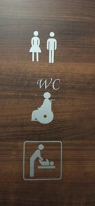Brązowe drzwi do toalety. Na nich pionowe oznakowanie za pomocą następujących piktogramów:na samej górze sylwetki kobiety i mężczyzny, poniżej litery WC,  pod nimi piktogram osoby na wózku, na samym dole piktogram osoby przewijającej dziecko. Wszystkie piktogramy srebrne.