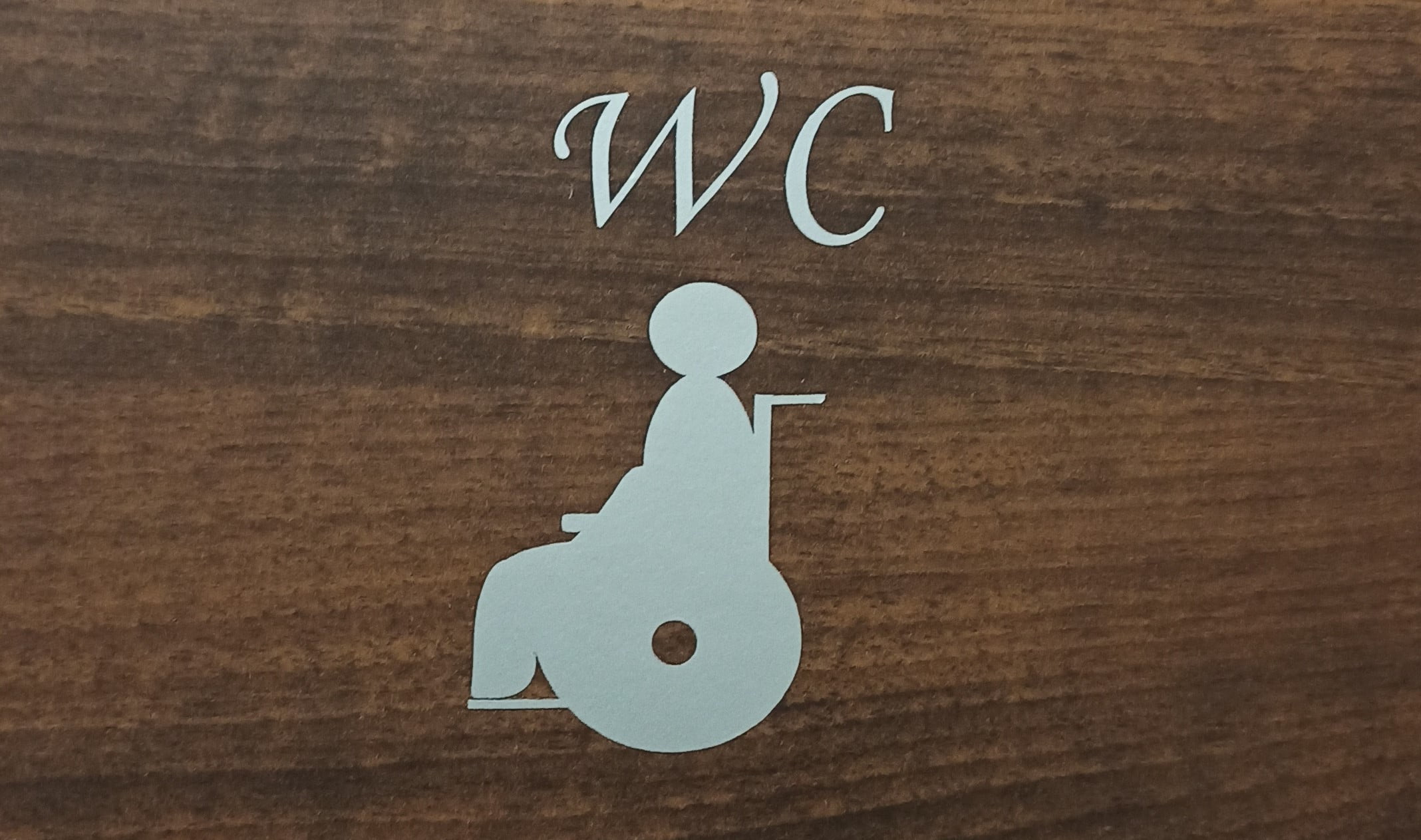 Na brązowym tle srebrny napis WC pod nim srebrny piktogram osoby na wózku