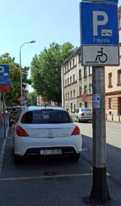 Miejsca parkingowe równoległe wzdłuż chodnika w paśmie ulicy. Najbliższe osobie robiącej zdjęcie miejsce parkingowe dedykowane jest osobom z niepełnosprawnością – na latarni przed miejscem zarówno niebieska tabliczka ze znakiem P i informacją o tym, że jest jedno takie miejsce, jak i biała tabliczka z piktogramem osoby na wózku. Samo miejsce nie różni się ani nawierzchnią ani rozmiarem od otaczających je miejsc.