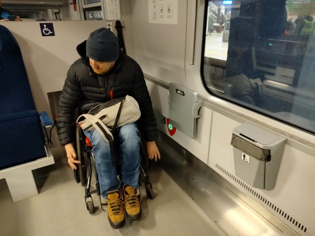 Mężczyzna na wózku parkuje na miejscu dedykowanym osobom na wózku w pociągu. Miejsce pozbawione jest miejsca siedzącego i oznaczone piktogramem osoby na wózku. W bezpośrednim sąsiedztwie miejsca przycisk przyzywowy zamontowany na wysokości około 40 – 50 cm – jest łatwo dostępny i podświetlony na zielono.