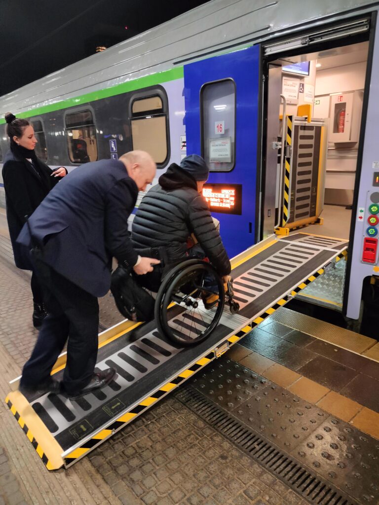 Peron na dworcu kolejowym. Przy nim pociąg z otwartymi drzwiami, między wejściem do wagonu i peronem rozłożona rampa przenośna. Konduktor pomaga pokonać rampę mężczyźnie na wózku.