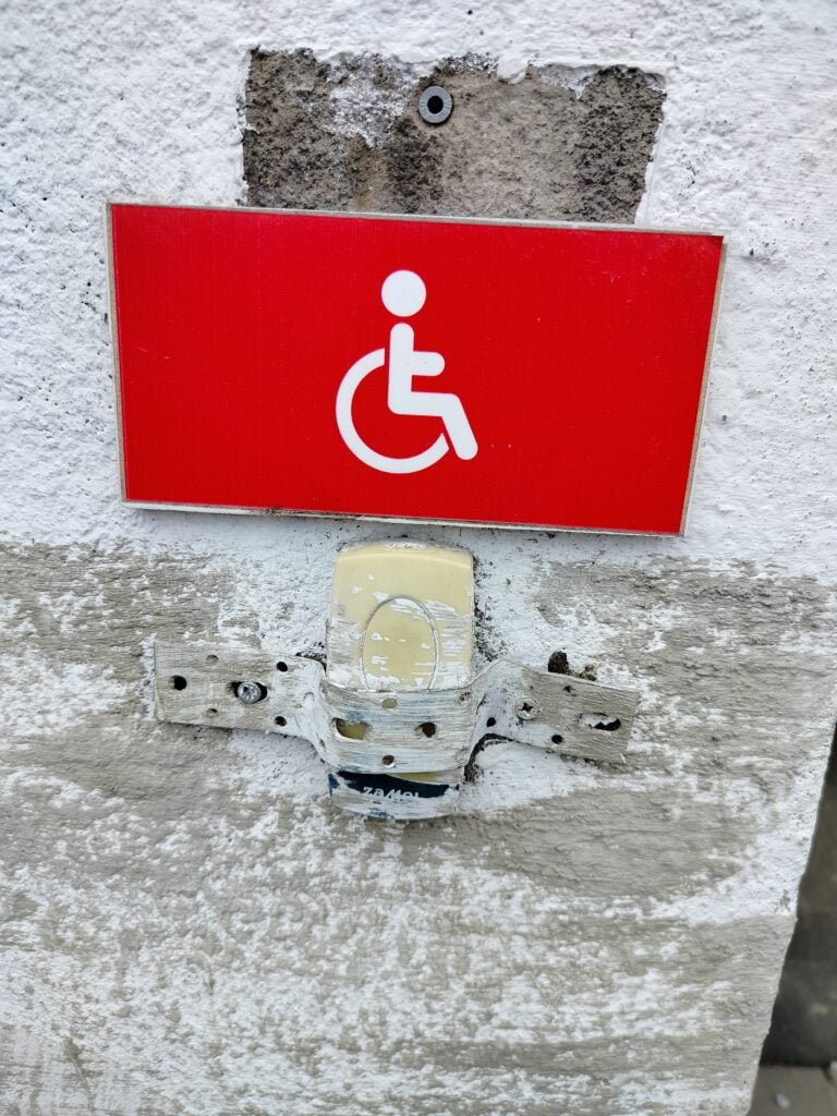 Zbliżenie na fragment ściany. Na niej czerwona tabliczka z białym piktogramem osoby na wózku. Pod tabliczką żółty plastikowy przycisk, w żółtym plastikowym obrabowaniu. Przycisk częściowo pomalowany biała farbą, dodatkowo umieszczony jest w metalowych objęciu, które uniemożliwia wciśnięcie przycisku.