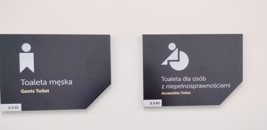 Jasno beżowa ściana, na niej sąsiadujące w poziomie ze sobą czarne prostokątne tablice z obciętym prawym dolnym rogu. W lewym dolnym rogu na jasnym tle numerek – w lewy dolnym rogu tabliczek na jasnym tle numer: na lewej 1.3.01, na prawej 2.3.03. Lewa tabliczka ma na sobie jasną postać męską (nieintuicyjną) z podpisem Toaleta damska i Gents Toilet. Prawa tabliczka z nieintuicyjnym symbolem osoby na wózku i podpisem Toaleta dla osób z niepełnosprawnościami Accessible Toilet.