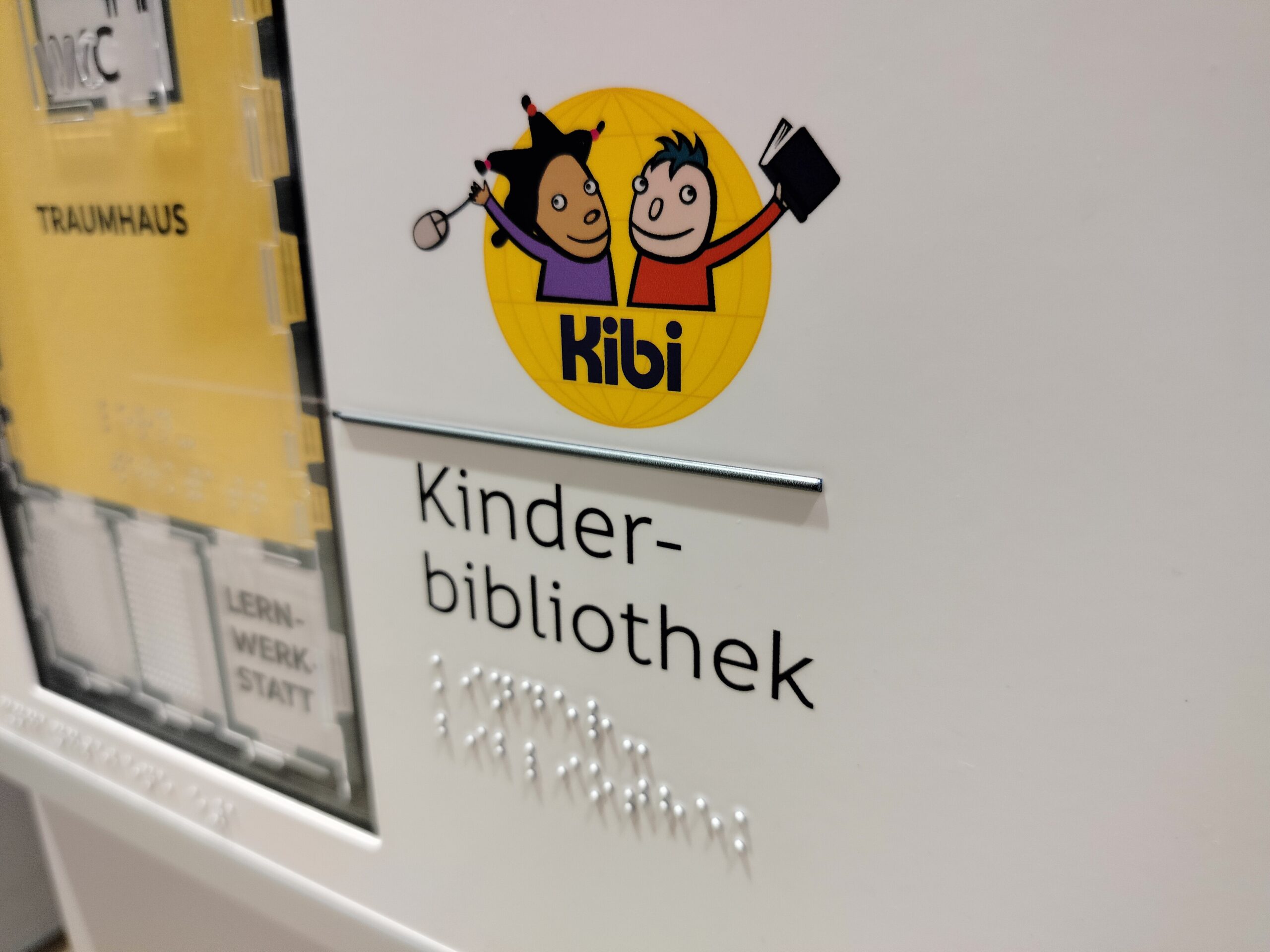 widoczny kawałek planu tyflograficznego, nam im logo Kibi - dwoje dzieci zwróconych do siebie - widoczne są od pasa w górę. Po prawej stronie chłopiec w czerwonej bluzce z długim rękawem, jasna twarz, krótkie nastroszone czarne włosy, w lewej podniesionej do góry ręce czarno oprawiona książka. Po lewej stronie dziewczynka w fioletowej bluzce z długim rękawem, ciemna twarz, długie czarne włosy zebrane w 4 kucyki, w podniesionej do góry prawej ręce myszka do komputera, całość na żółtym przekroju kuli imitującej kulę ziemską. Pod dziećmi, na tle kulo czarny napis Kibi. Poniżej napis Kinder bibliothek czarnymi literami i w alfabecie Braille'a.