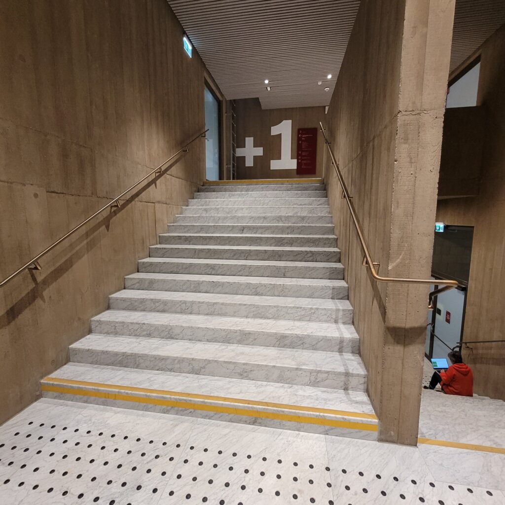 Ciąg schodów - przed nim pola uwagi, pierwszy i ostatni stopień oznaczony kontrastem koloru i faktury, na górze widoczne bardzo duże oznaczenie poziomu +1 - biała czcionka na czarnym tle.