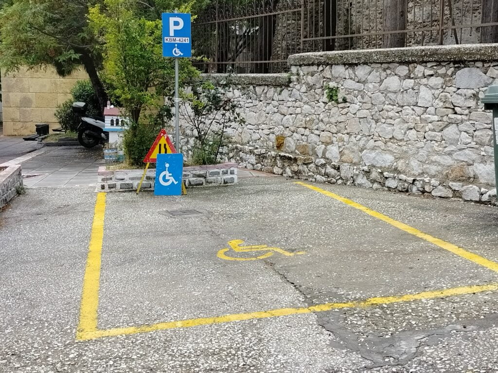 Zaznaczone żółtą obwódką miejsce parkingowe oznakowane również żółtym piktogramem osoby na wózku. Dodatkowo oznakowanie pionowe znakiem P i piktogramem osoby na wózku. Litera i symbol białe na niebieskim tle. Na krańcu miejsca stoi znak oznaczający zwężenie drogi a na nim oparty biały piktogram osoby na wózku na niebieskim tle.