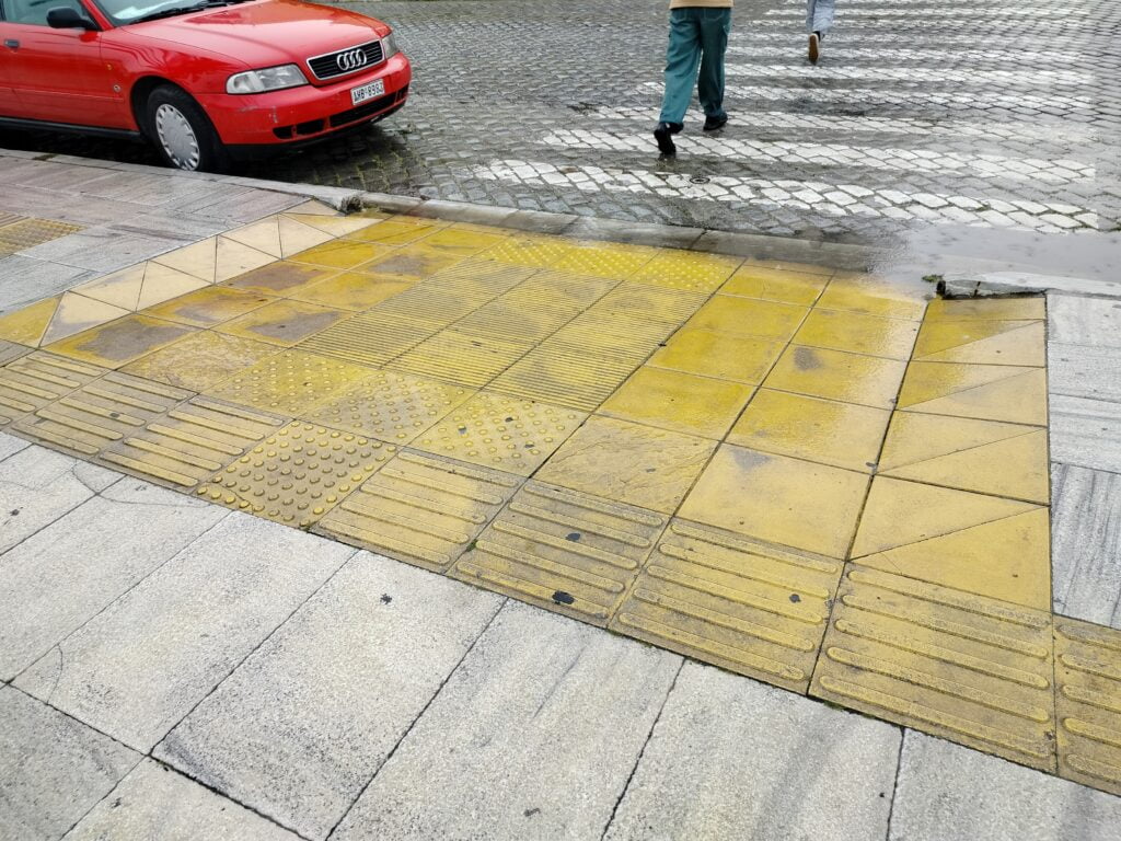 Przejście dla pieszych z obniżonym krawężnikiem. Całość obniżenia wyłożona żółtymi płytkami w różne wzory. 4 pola uwagi na styku ulicy i chodnika, od strony chodnika pojedyncza płytka w pierwszym rzędzie oraz 4 w drugim. Pierwszy rzad płytek od strony chodnika to ścieżka prowadząca. Pozostałe płytki w ramach obszaru obniżenia to żółte kwadraty bez żadnej faktury. Obramowanie boczne obniżenia to pasy ułożone z par żółtych płytek trójkątnych.
