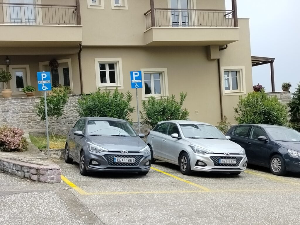 Widok na budynek i usytuowany przed nim parking. Na parkingu widoczne 2,5 miejsca - wszystkie zajęte. Po lewej stronie 2 miejsca parkingowe dedykowane osobom z niepełnosprawnością oznakowane żółtą obwódką i pionowym oznaczenie białą literą P i białym symbolem osoby na wózku na niebieskiej tablicy.