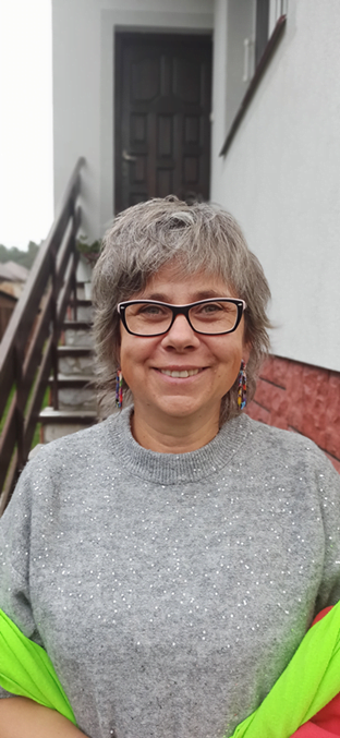 Małgorzata Peretiatkowicz na tle schodów prowadzących do budynku. Kobieta koło pięćdziesiątki, szpakowate, ktrótkiei włosy, na nosie okulary.