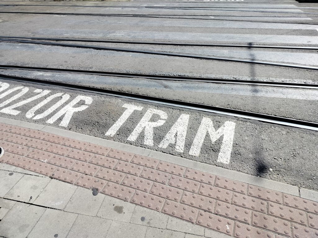 Styk ulicy z torami tramwajowymi i chodnika. Na chodniku tuż przed jezdnią ciąg 4 cienkich czerwonych płytek z wypustkami. Na jezdni napis białą farbą POZOR TRAM.