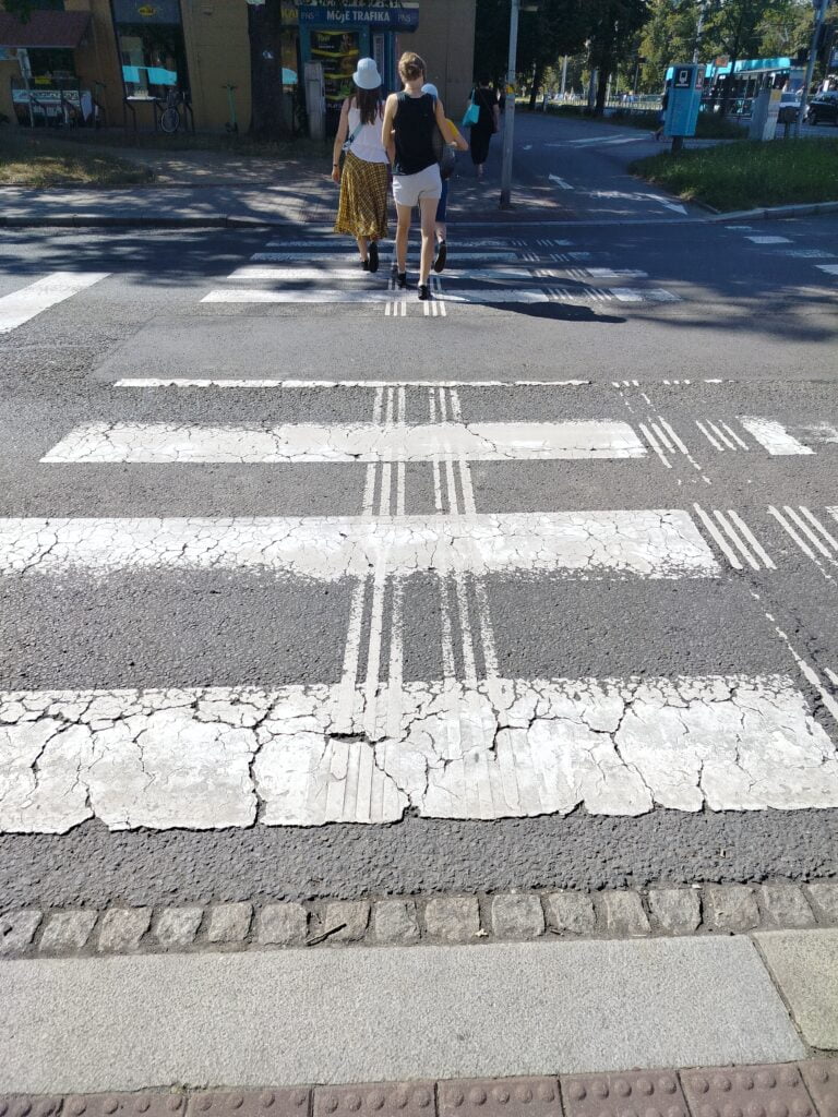 Widok z góry na przejście dla pieszych. pasy poziome w wielu miejscach starte. Dodatkowo pojawiają się pasy pionowe również miejscami zdarte. Na końcu przejścia tyłem idą 2 osoby.