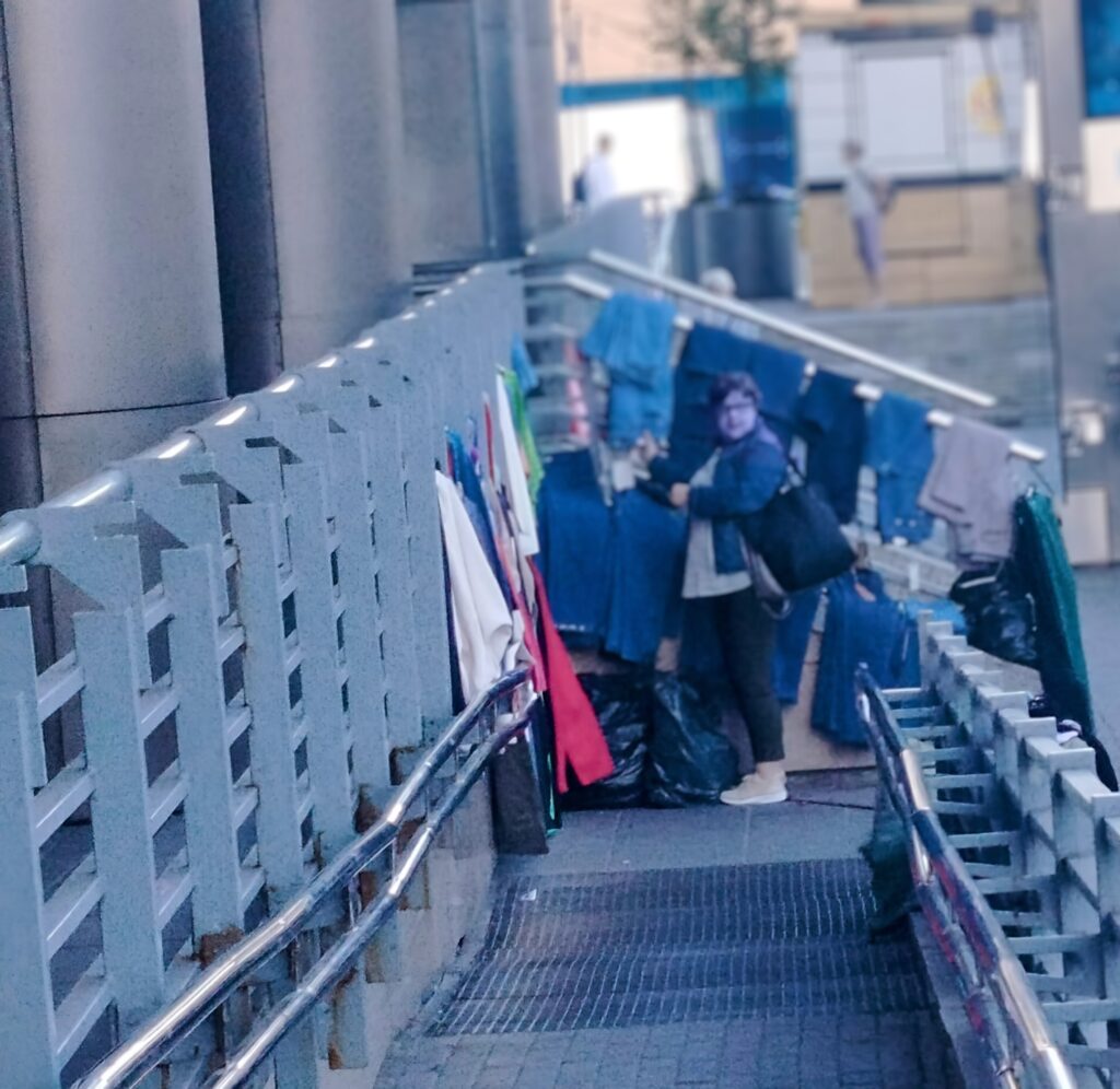 Widok na pochylnię z góry. Na jej dole rozmyta kobieta trzyma w ręku wieszak z bluzką, który właśnie zdjęła. Kobieta stoi na pochylni,a wieszaki z ubraniami wiszą wokół niej.