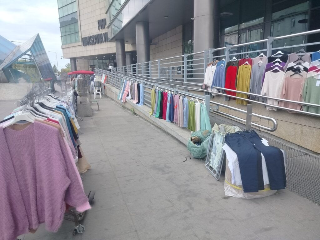 Widok na pochylnię z przodu na jej pochwytach od strony chodnik wiszą kolorowe bluzki i spodnie na plastikowych wieszakach. Z przodu kadru wieszak sklepowy na wieszaki a na nich kolejne ubrania.